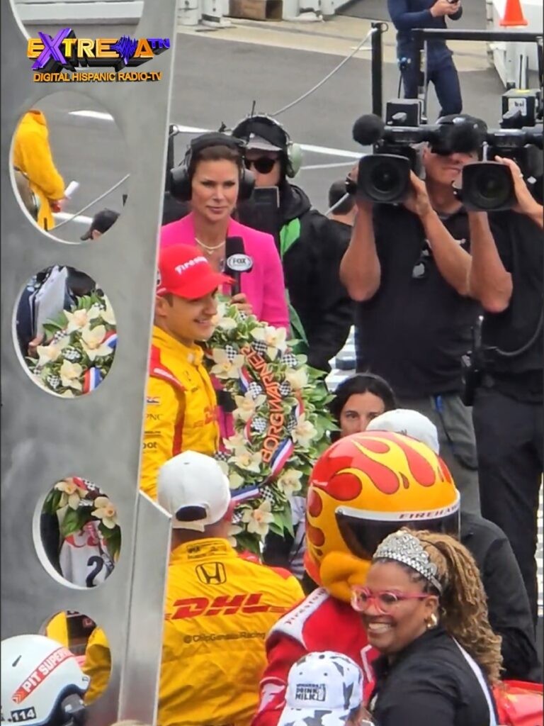 Álex Palou con corona de flores en la ceremonia de victoria de la Indy 500 2025 rodeado de medios y cámaras en el Indianapolis Motor Speedway