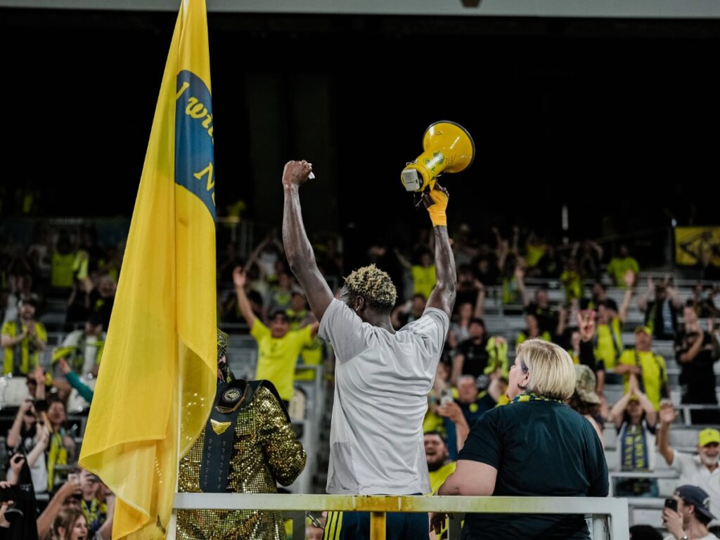 Maxwell Woledzi sostiene un megáfono amarillo liderando los cánticos con los aficionados